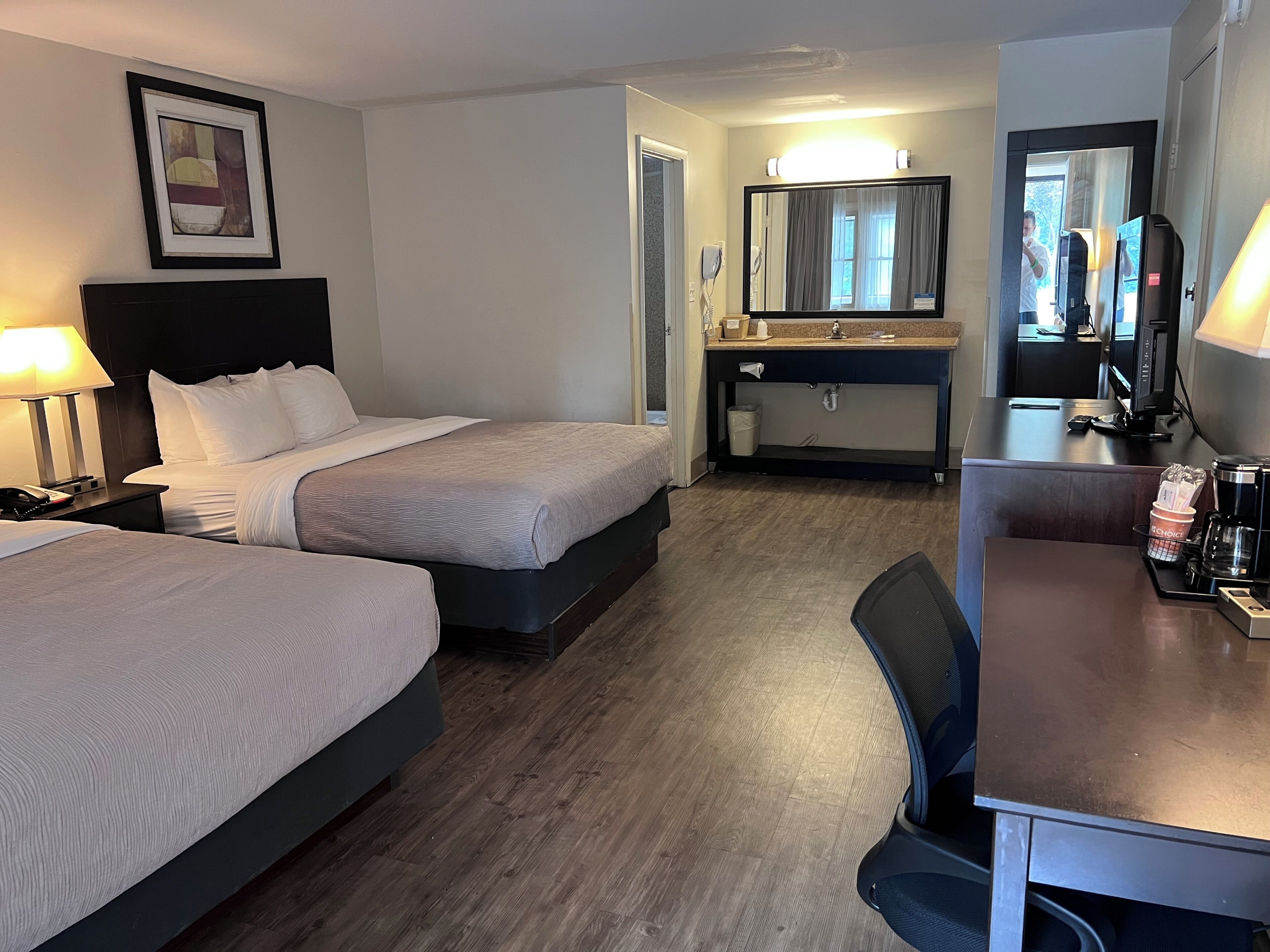 Habitación de hotel con dos camas, un escritorio, un espejo y un baño visible a través de una puerta abierta.
