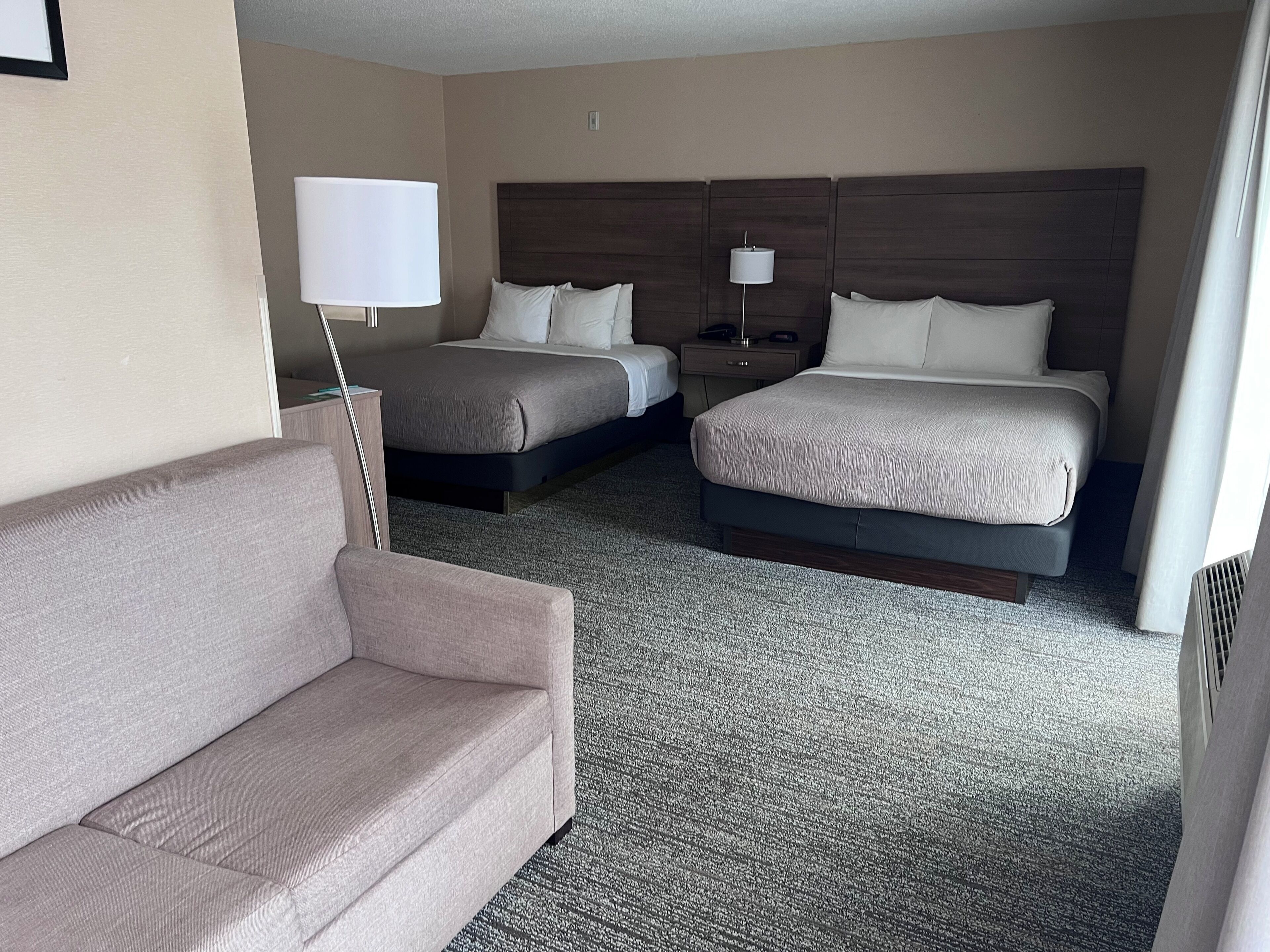 A hotel room with a sofa, two beds, a nightstand, and a lamp.