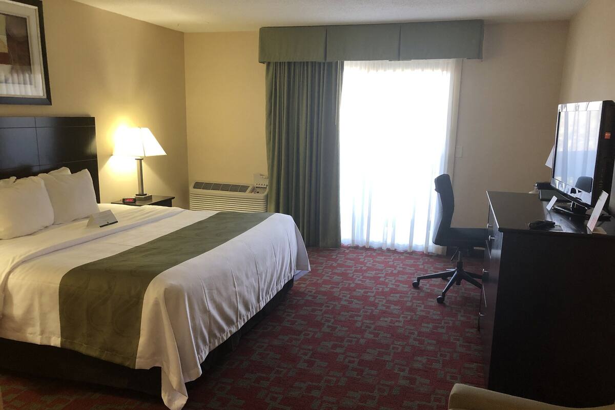 Ein Hotelzimmer mit einem großen Bett, einem Schreibtisch mit Computer, einem Stuhl, einer Lampe und einem fenster mit Vorhängen.