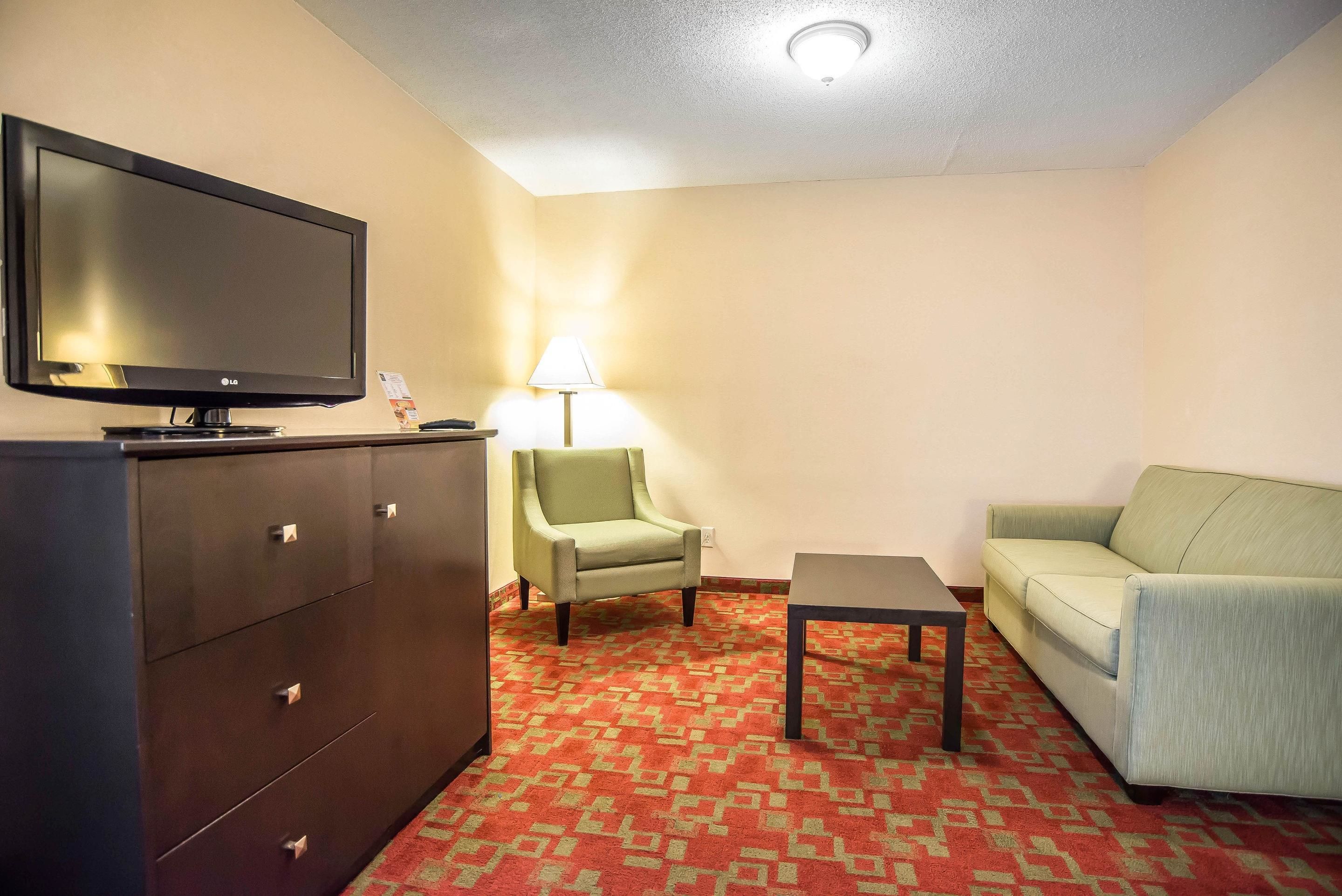 A hotel room with a flat-screen TV, a green armchair, a light-colored sofa, a small table, and a patterned carpet.