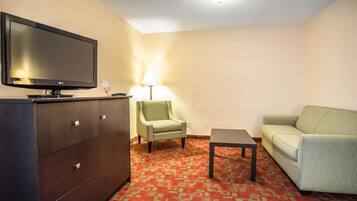 A hotel room with a flat-screen TV, a green armchair, a light-colored sofa, a small table, and a patterned carpet.