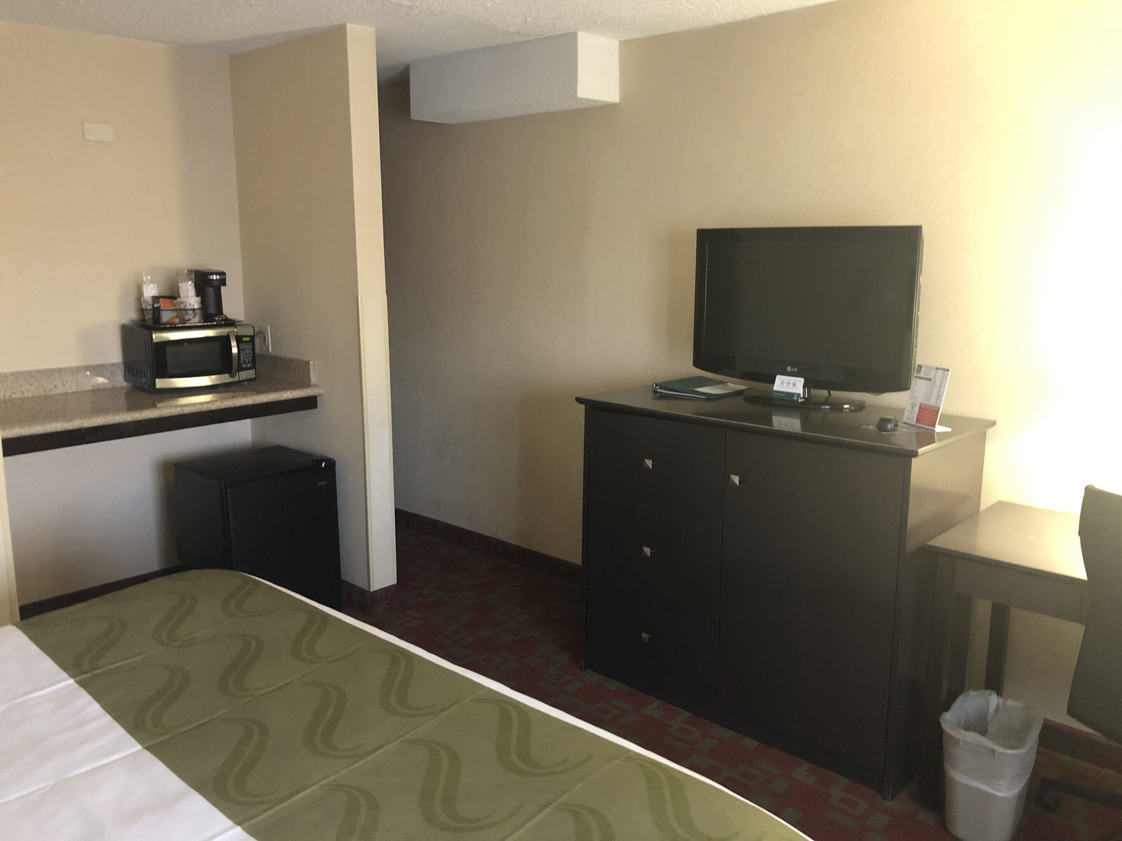 A hotel room with a bed, a desk with a microwave, a television, and a small trash bin.