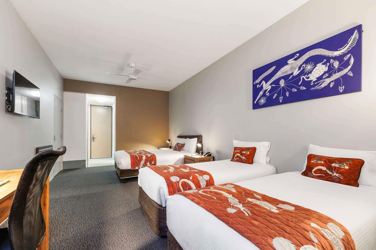 Ein Hotelzimmer mit zwei Betten, einem Schreibtisch, einem Stuhl, einem Deckenventilator, einem Flachbildfernseher und einem großen Gemälde an der Wand.