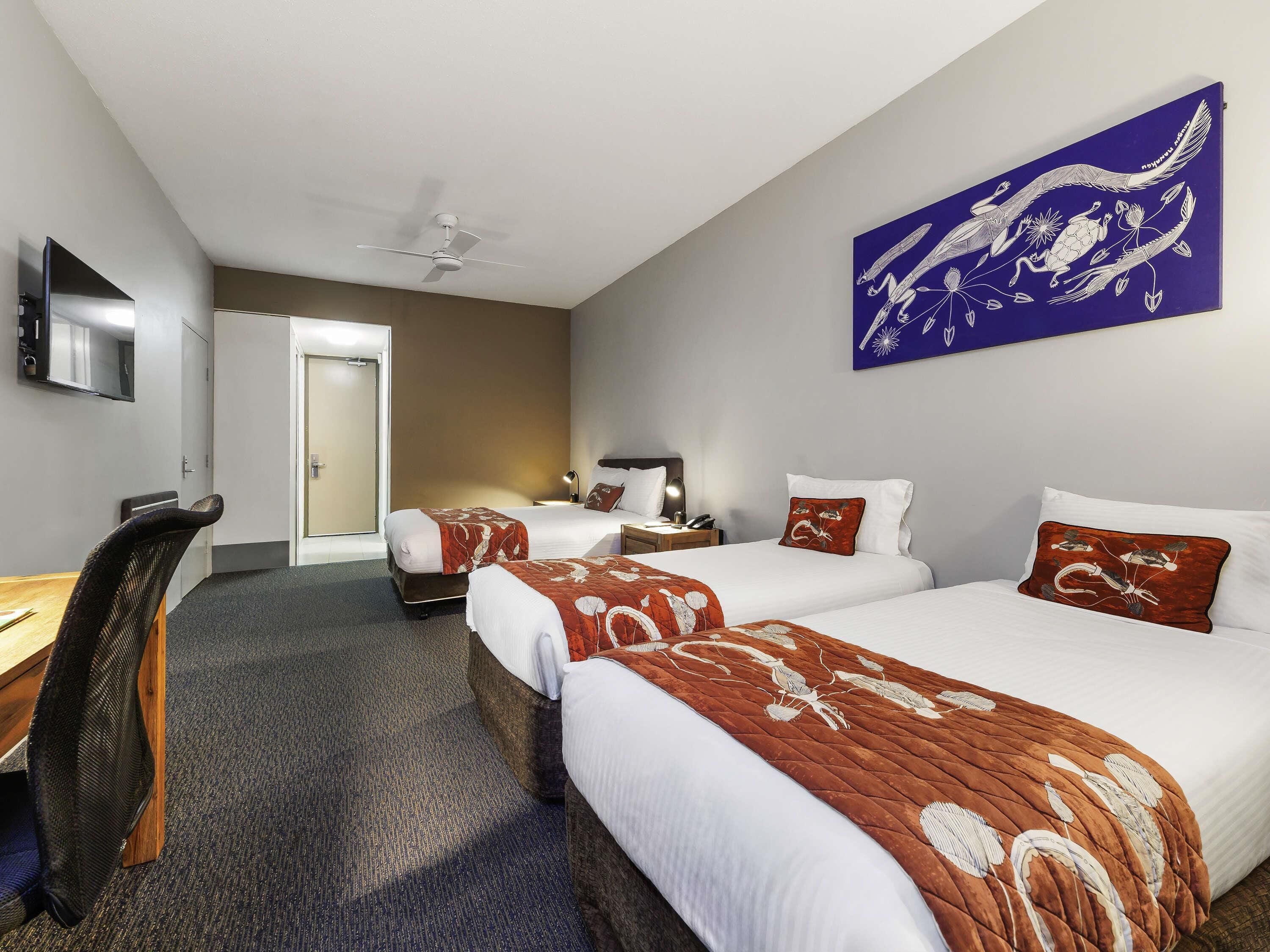 Ein Hotelzimmer mit zwei Betten, einem Schreibtisch, einem Stuhl, einem Deckenventilator und einem Bild an der Wand.