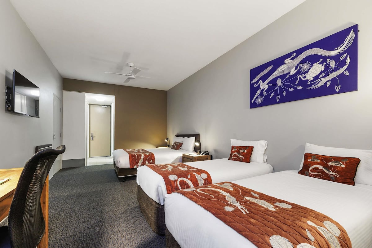 Ein Hotelzimmer mit zwei Betten, einem Schreibtisch, einem Stuhl, einem Deckenventilator und einem Bild an der Wand.