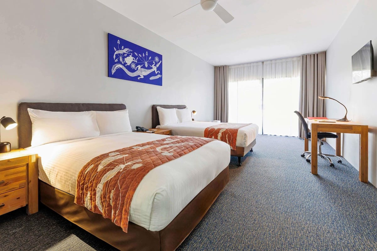 Ein Hotelzimmer mit zwei Betten, einem Schreibtisch, einem Stuhl, einem Deckenventilator und einem großen Fenster mit Vorhängen.