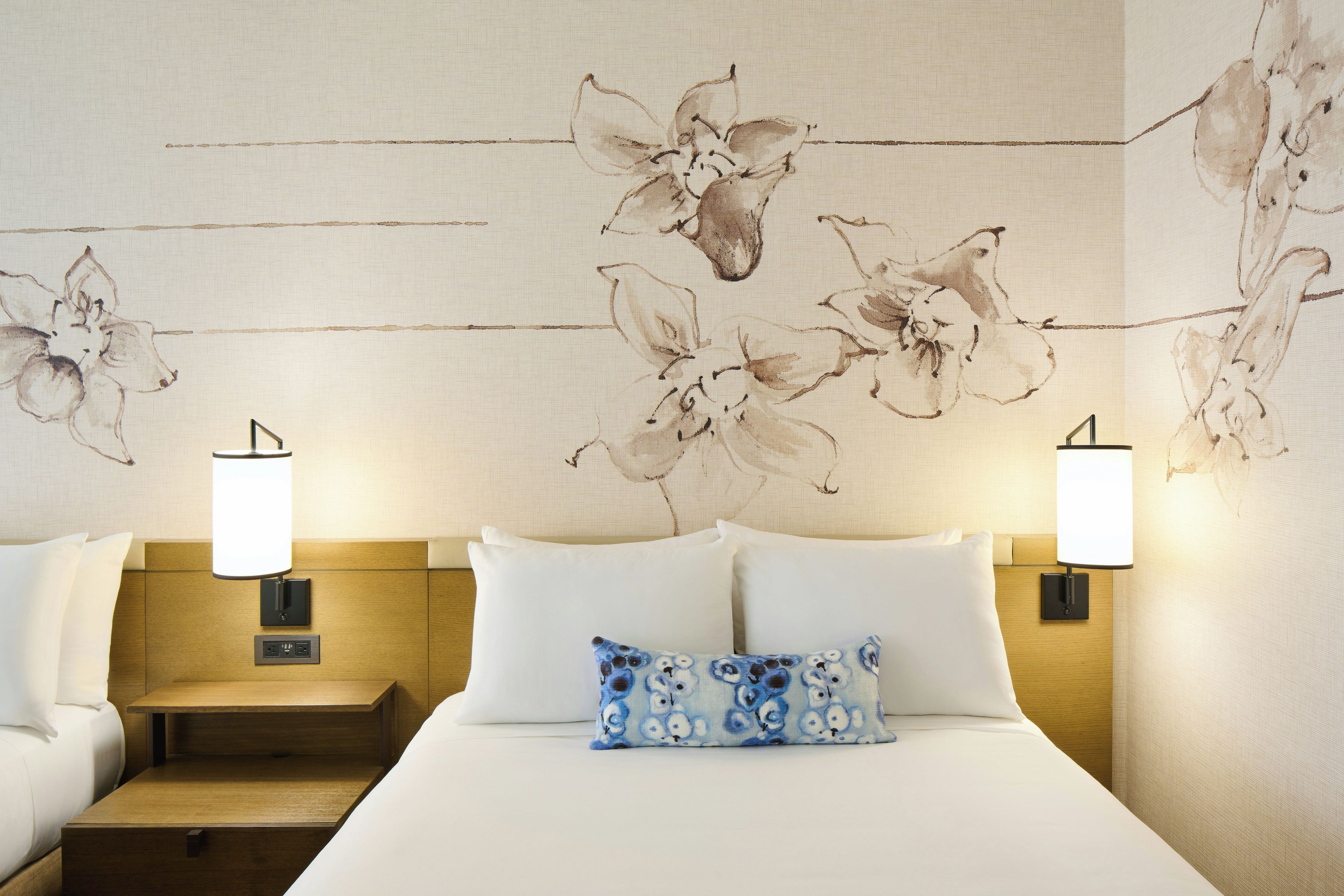 A hotel room with a bed, two bedside lamps, a nightstand, and a wall with floral artwork.