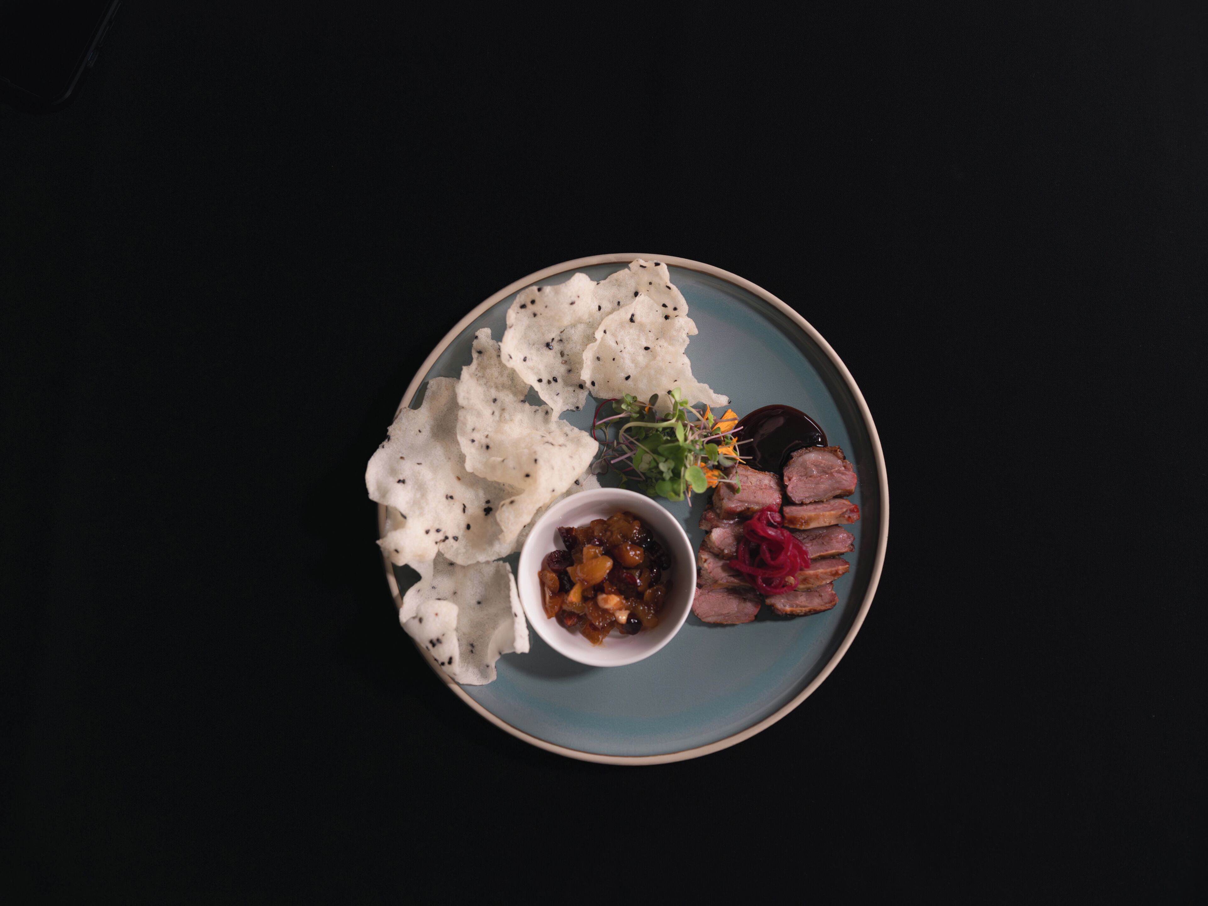 Un plato con carne en rodajas, verduras encurtidas y un recipiente pequeño con salsa sobre un fondo oscuro.