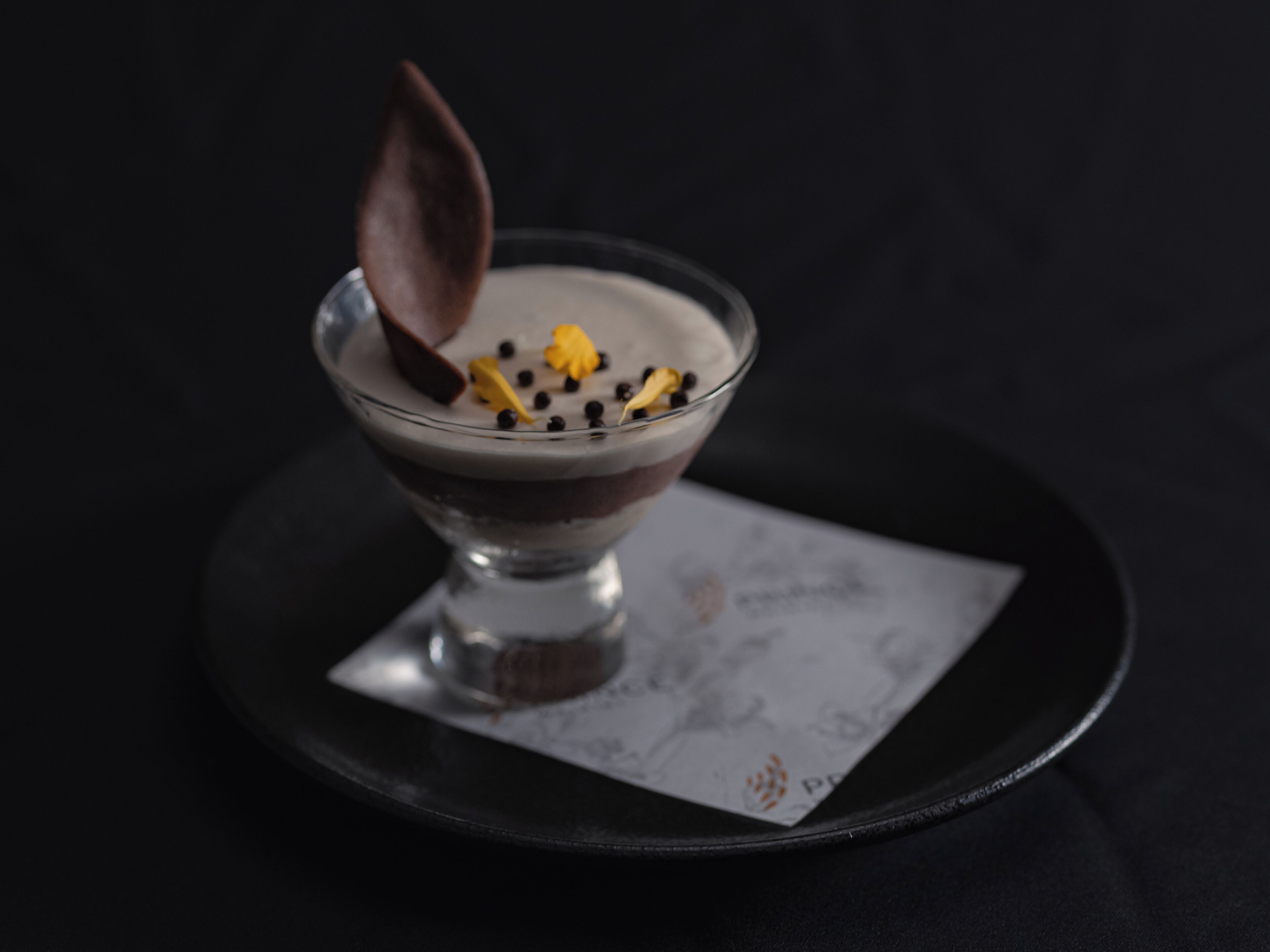 A dessert in a glass bowl on a black plate with a napkin underneath.