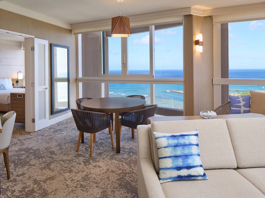 Habitación de hotel con un amplio ventanal que ofrece vistas al océano, una mesa redonda de comedor con sillas, un sofá con un cojín azul y una pequeña mesa auxiliar con una lámpara.