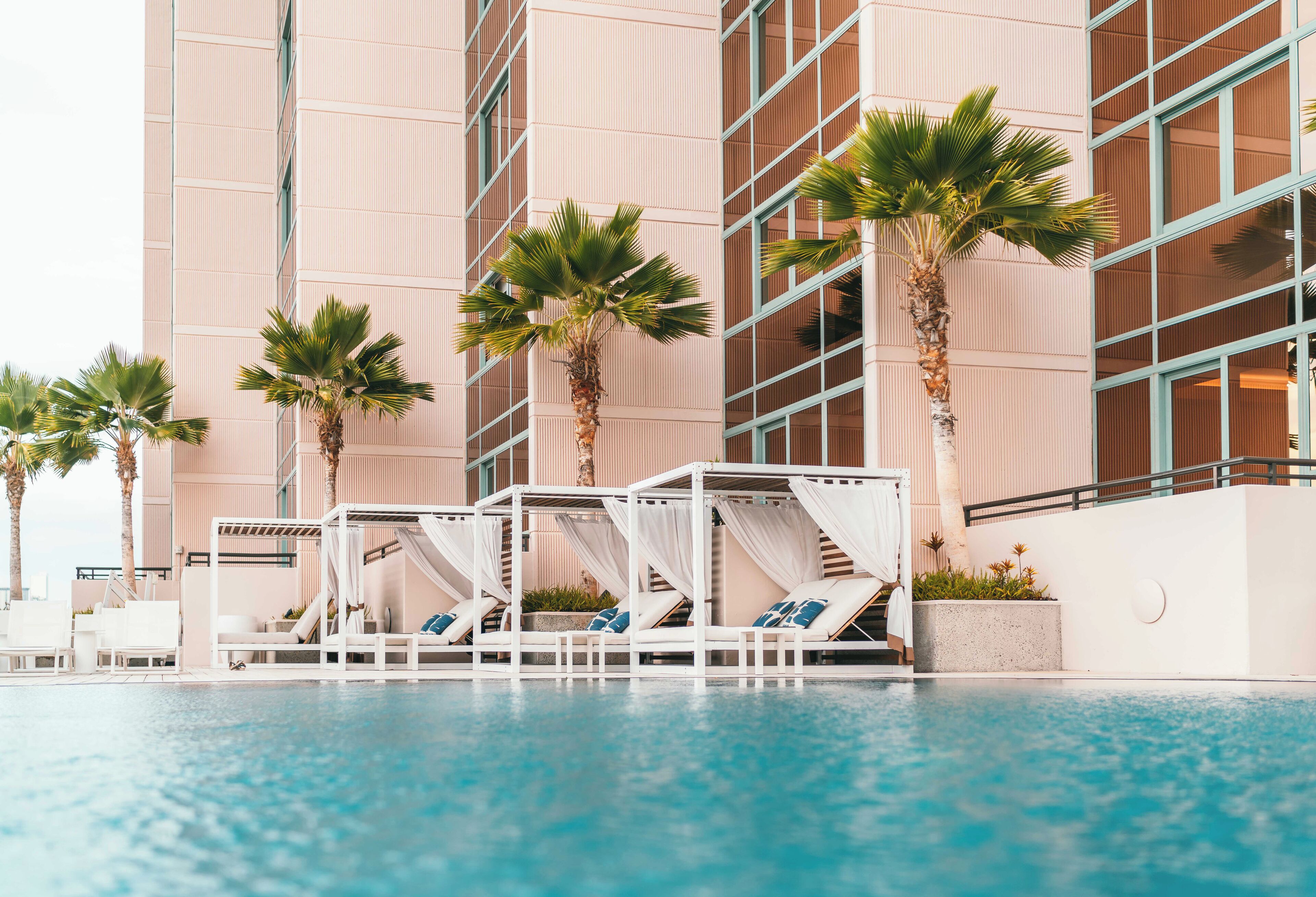 A pool area with lounge chairs and palm trees, surrounded by a modern building with large windows.
