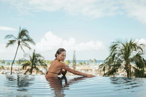 A woman in a bikini sitting by a pool with a beach and palm trees in the background. - Prince Waikiki (Honolulu)