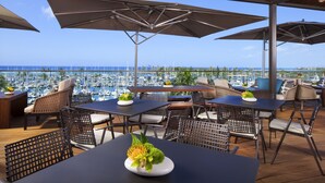 An outdoor dining area with tables and chairs, umbrellas, and a view of the marina.