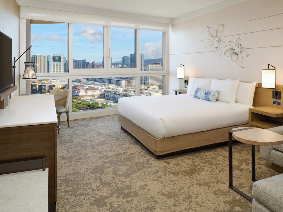 A hotel room with a large bed, a desk, a chair, a TV, and a view of the cityscape.