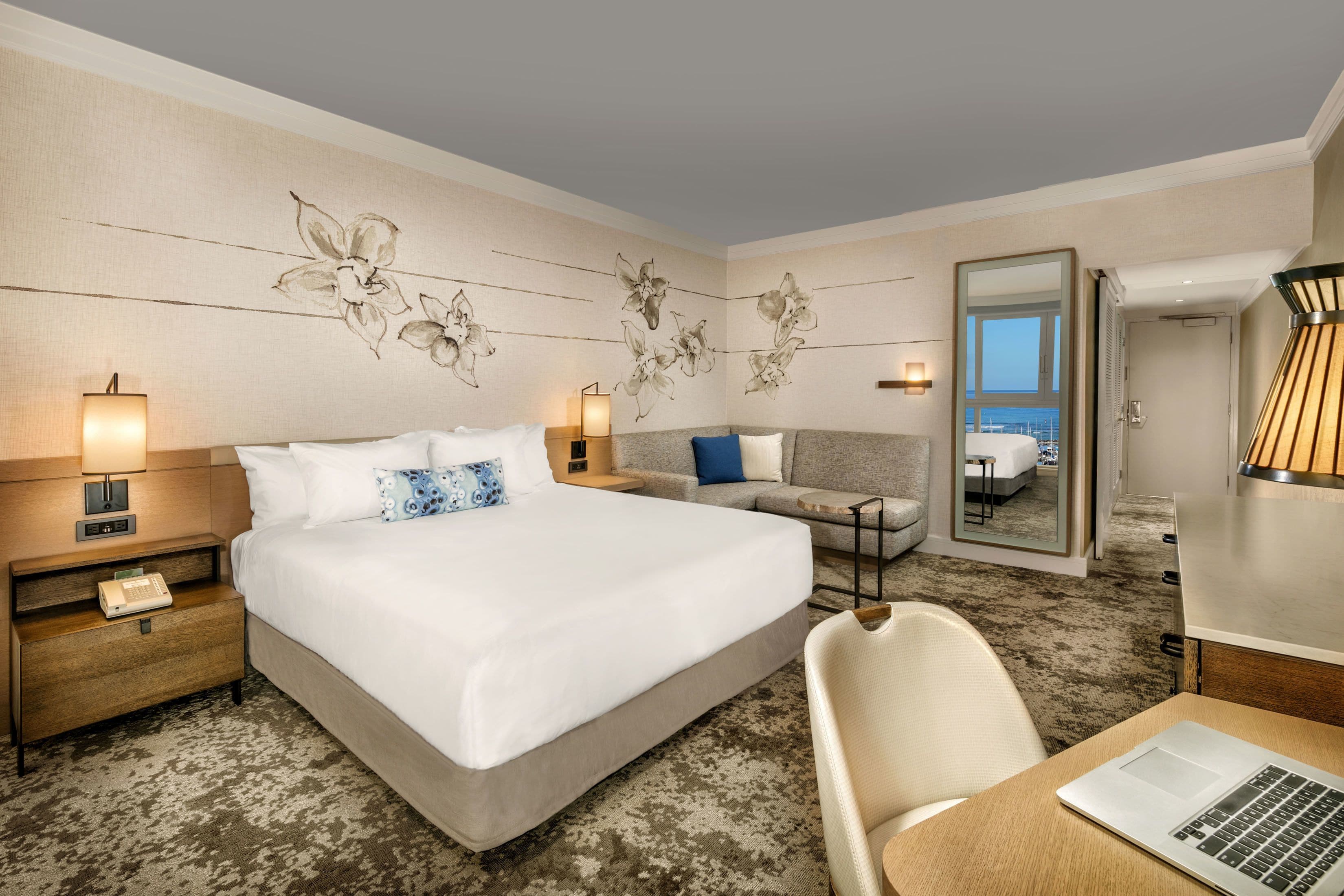Habitación de hotel con una cama grande, un escritorio con una computadora portátil, un sofá y un espejo que refleja un balcón.