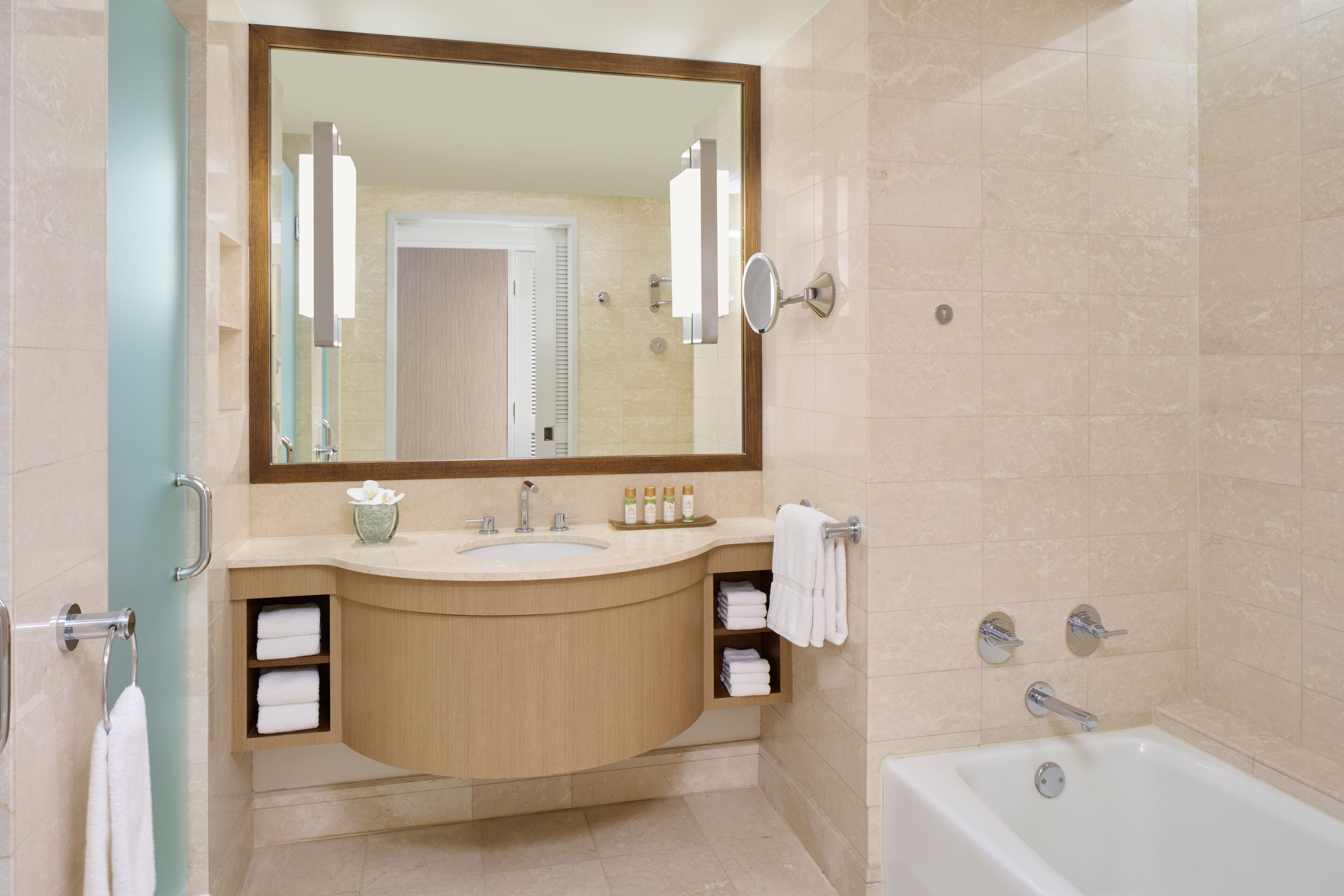 A modern bathroom with a large mirror, sink, and bathtub. There are towels and toiletries on the counter.