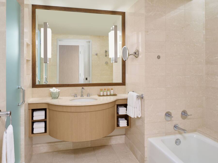 A modern bathroom with a large mirror, sink, and bathtub. There are towels and toiletries on the counter.