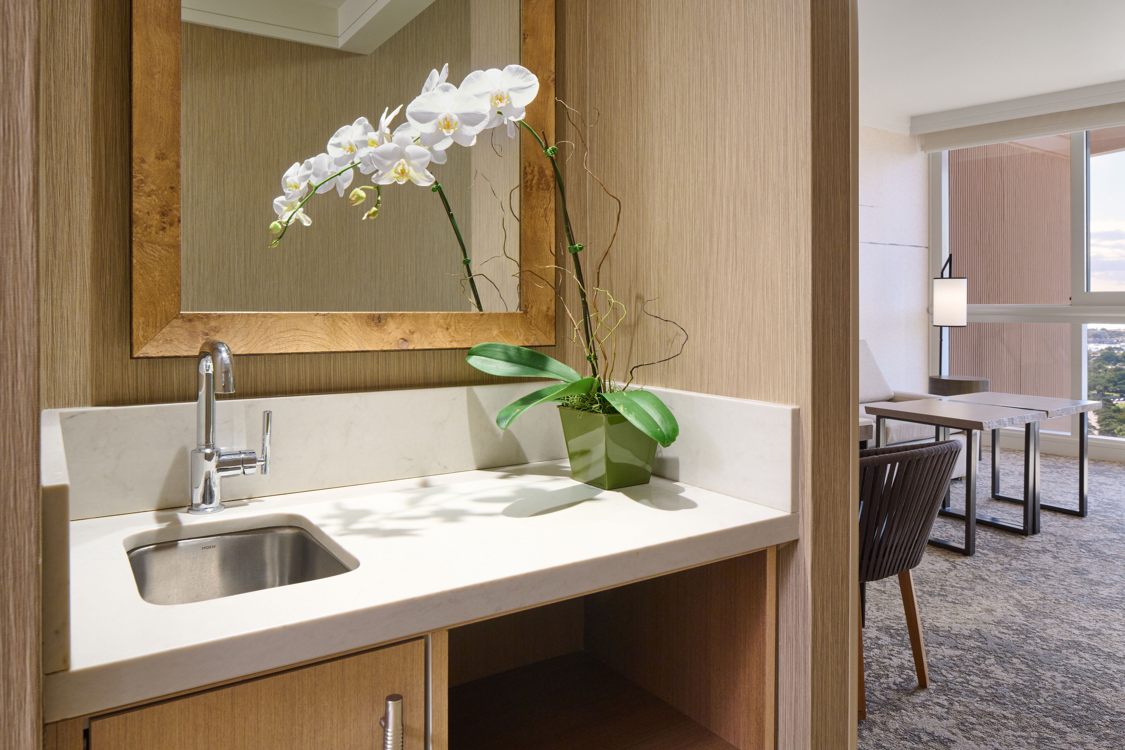 A modern bathroom with a wooden vanity, a sink, and a mirror. There is a potted orchid on the vanity.