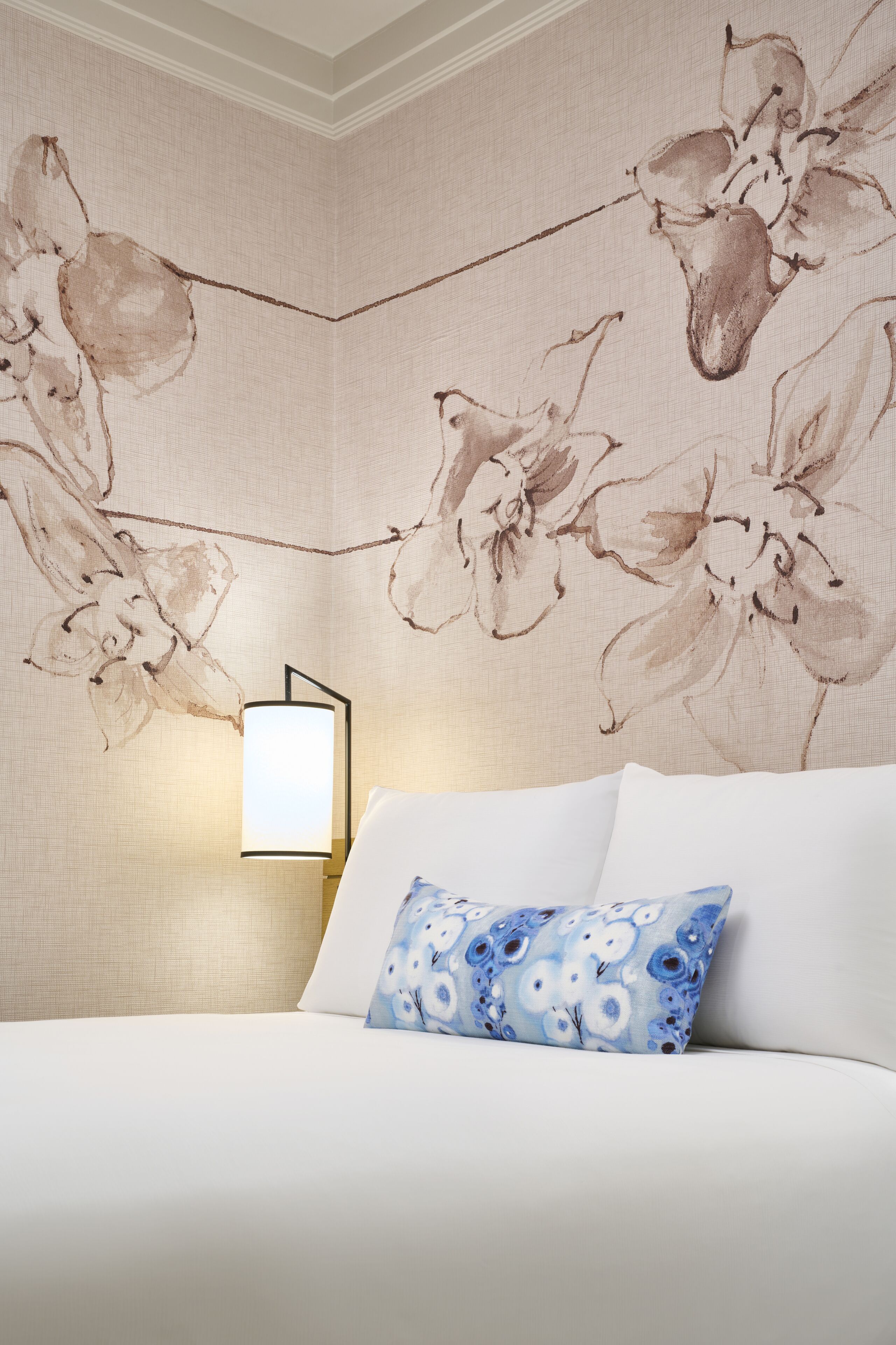 Quarto com papel de parede floral, luminária de cabeceira e travesseiro estampado.