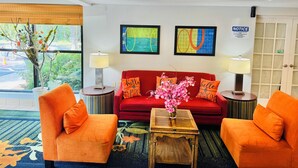 A hotel lobby with a red sofa, orange armchairs, a wooden coffee table, and two lamps. There are two framed artworks on the wall and a notice sign.