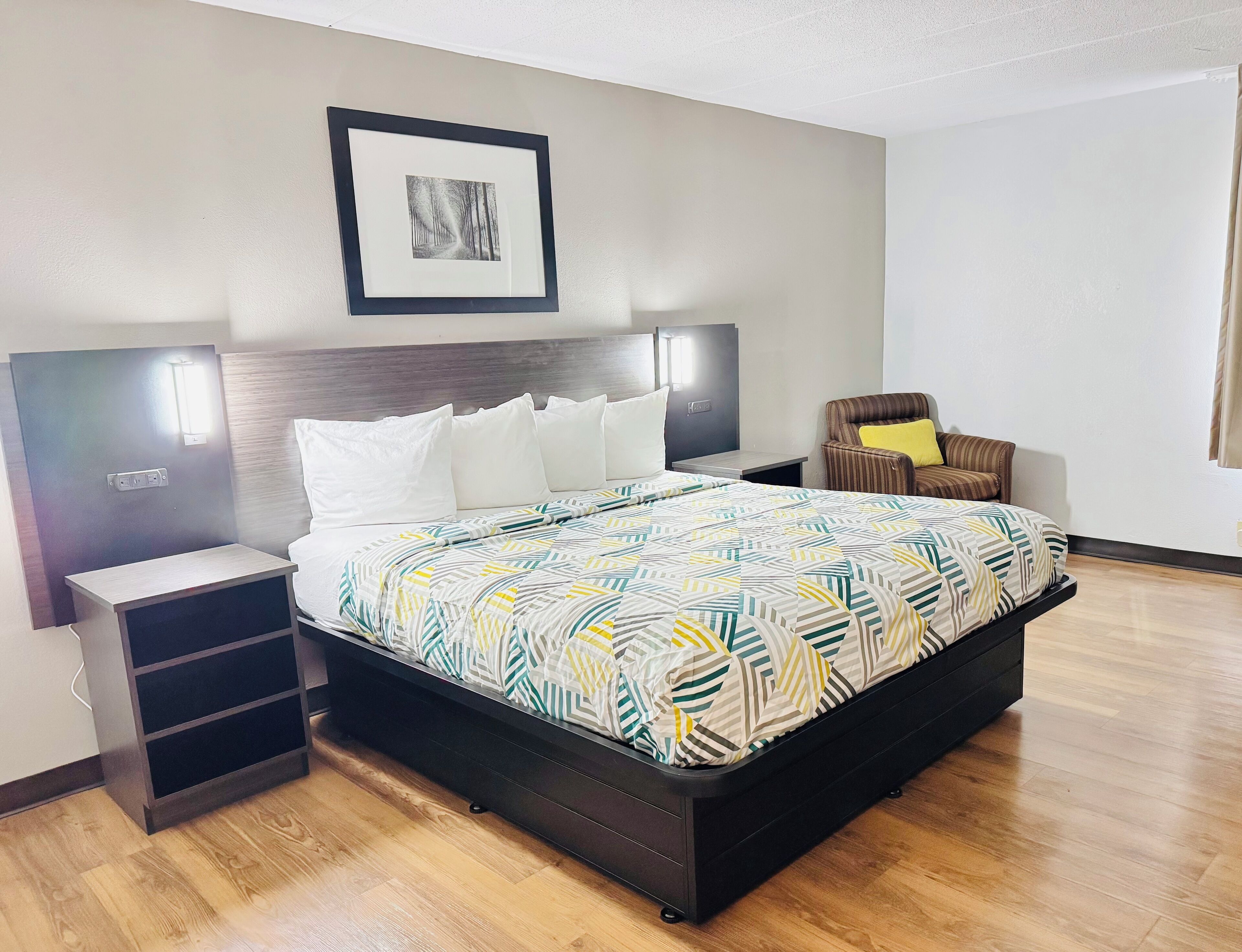A hotel room with a large bed, a nightstand, a chair, and a framed picture on the wall.