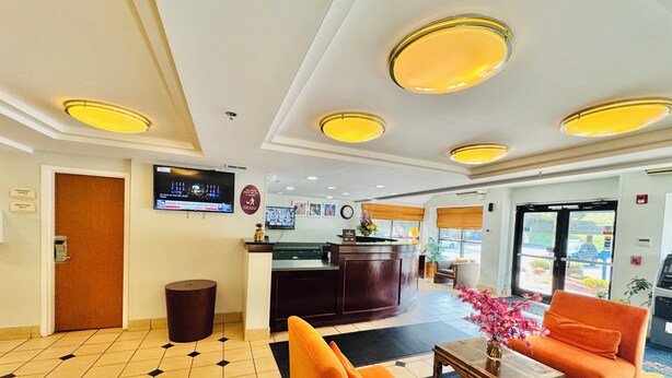 A hotel lobby with a reception desk, a television mounted on the wall, a trash can, and a seating area with orange chairs and a table.