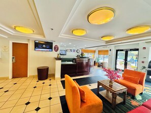 A hotel lobby with a reception desk, a television mounted on the wall, a trash can, and a seating area with orange chairs and a table. - Motel 6 Milford, CT (Milford)