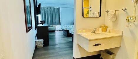 A hotel room with a wooden floor, a white sink, a mirror, and a bed with a blue and white patterned bedspread.