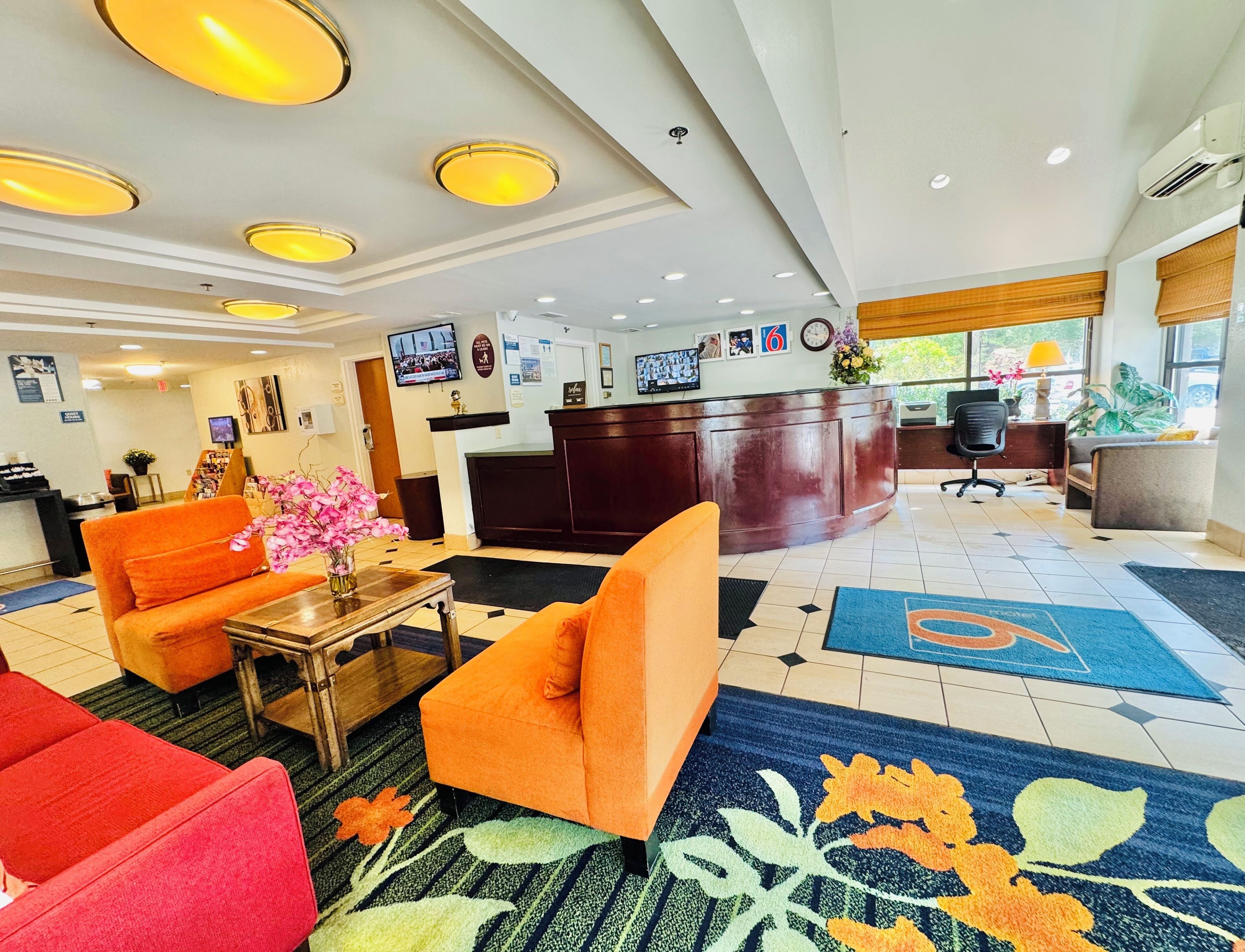 A hotel lobby with a reception desk, orange sofas, a coffee station, and a TV screen.