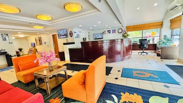 A hotel lobby with a reception desk, orange sofas, a coffee station, and a TV screen.