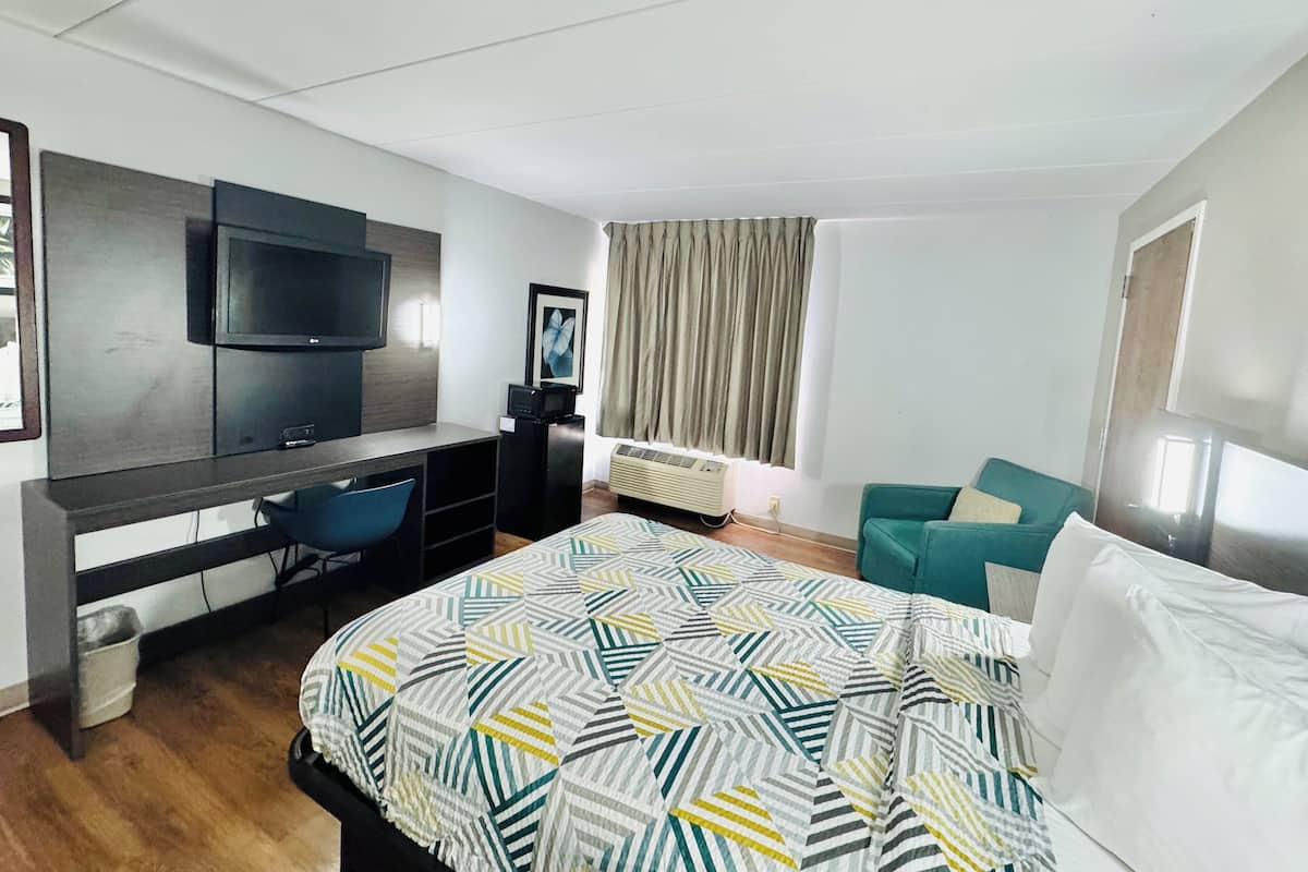 Ein modernes Hotelzimmer mit einem großen Bett, einem Schreibtisch mit Flachbildfernseher, einem bequemen, smaragdgrünen Sessel und einem großen Fenster mit Vorhängen.
