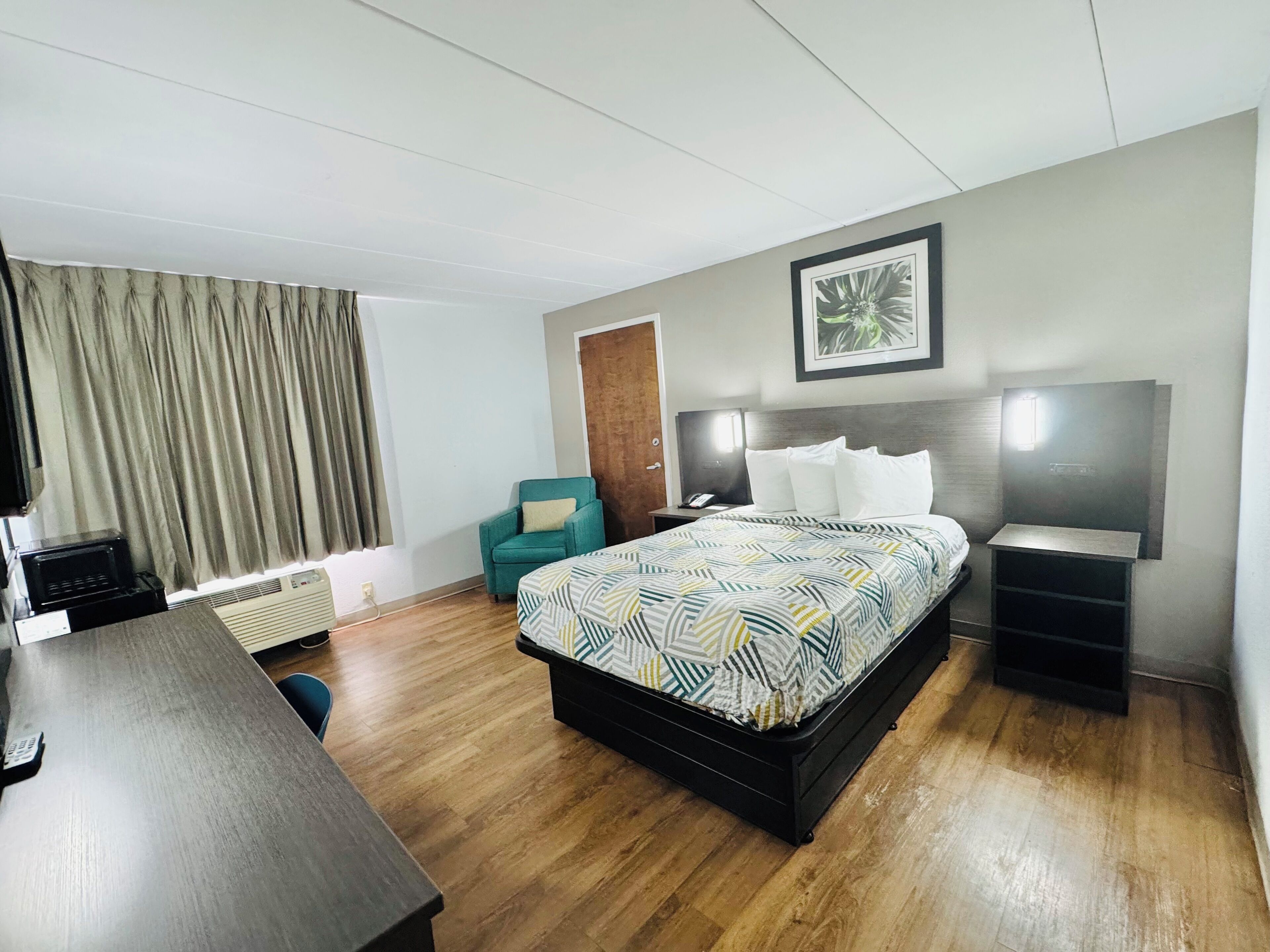 Habitación de hotel con cama, escritorio, silla, mesita de noche y televisor montado en la pared, además de una ventana con cortinas.