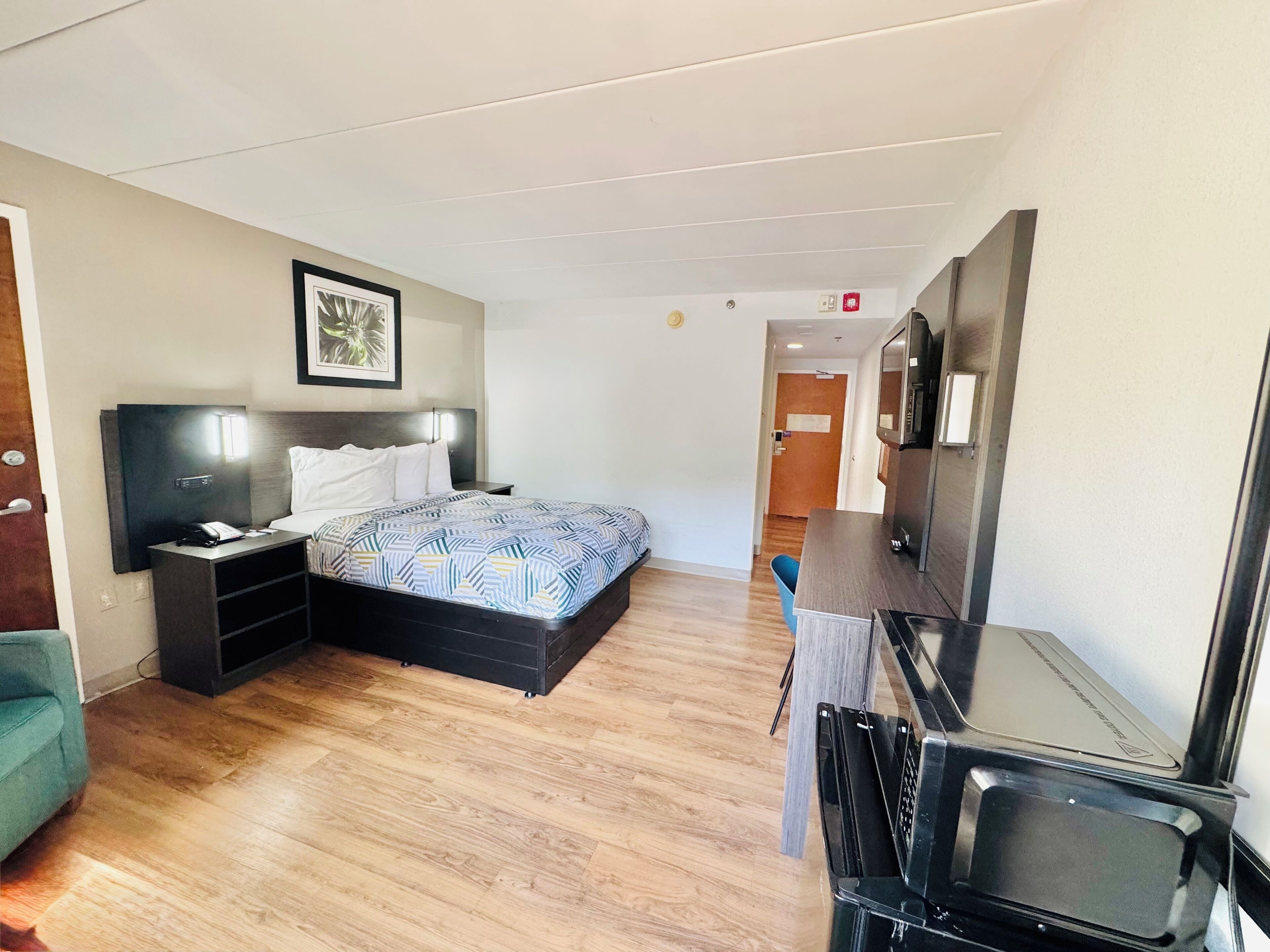 A hotel room with a bed, bedside table, and a desk with a chair. There is a door leading to another room and a wall-mounted TV.