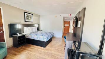 A hotel room with a bed, bedside table, and a desk with a chair. There is a door leading to another room and a wall-mounted TV.