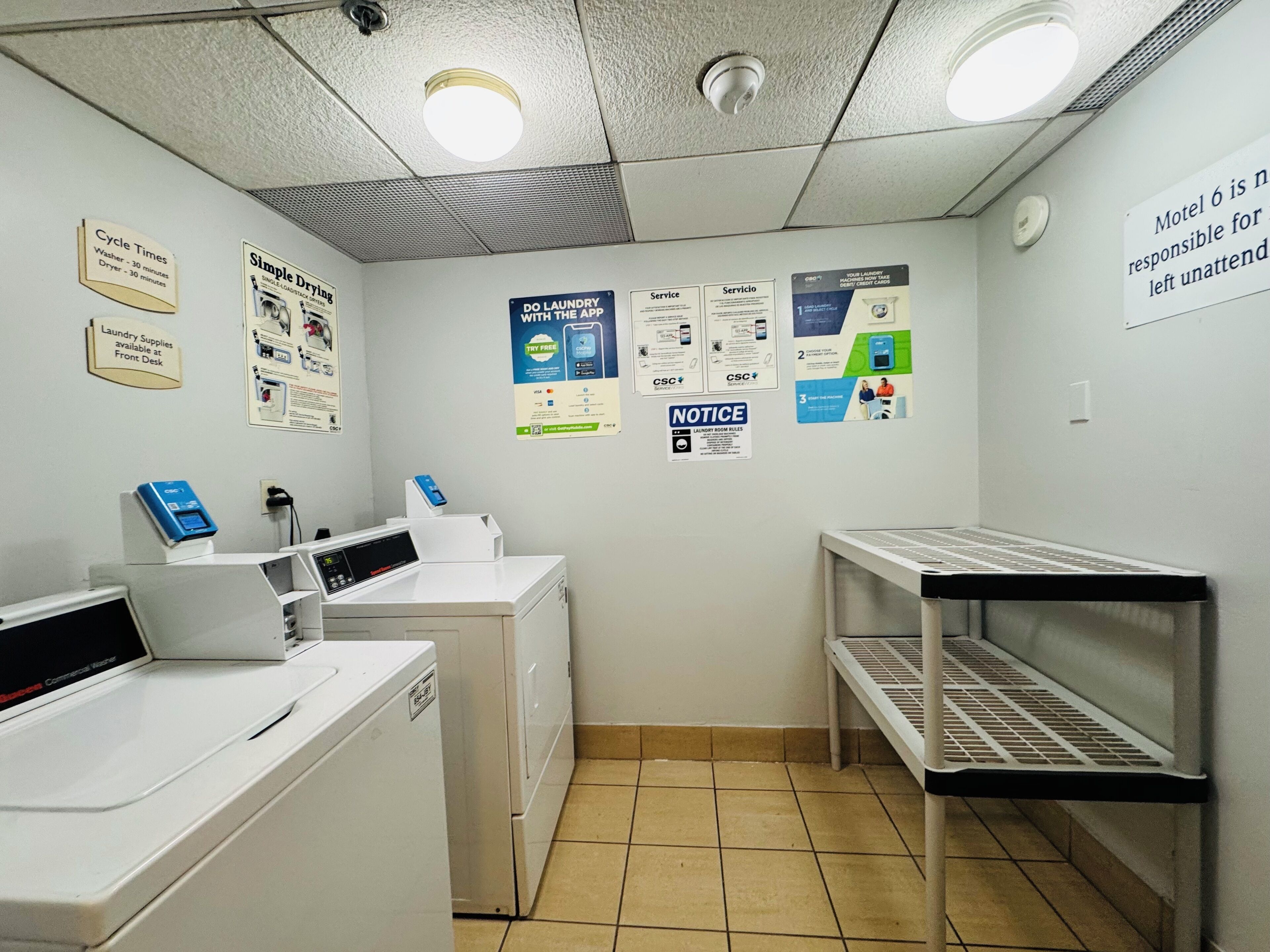 Une buanderie équipée d’une machine à laver industrielle, de séchoirs et d’une machine à laver en libre-service. Des panneaux indiquent la durée des cycles de lavage, les produits de lessive disponibles, et affichent également les règles d’utilisation de la buanderie.