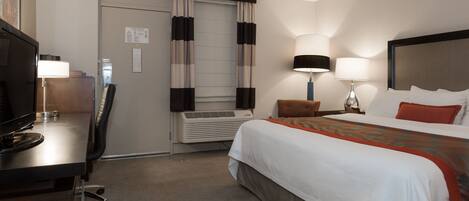 A hotel room with a bed, a desk with a television, a desk lamp, a wall-mounted TV, and a window with curtains.
