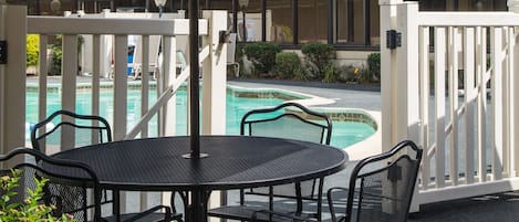 An outdoor patio with a round table and chairs, a large umbrella, and a pool in the background.