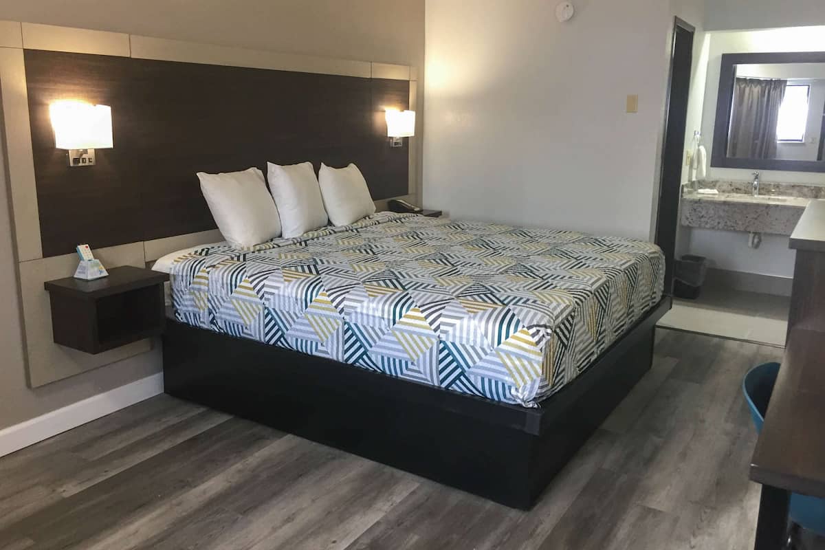 A modern hotel room with a large bed, a nightstand, a bedside lamp, and a bathroom with a mirror and sink.