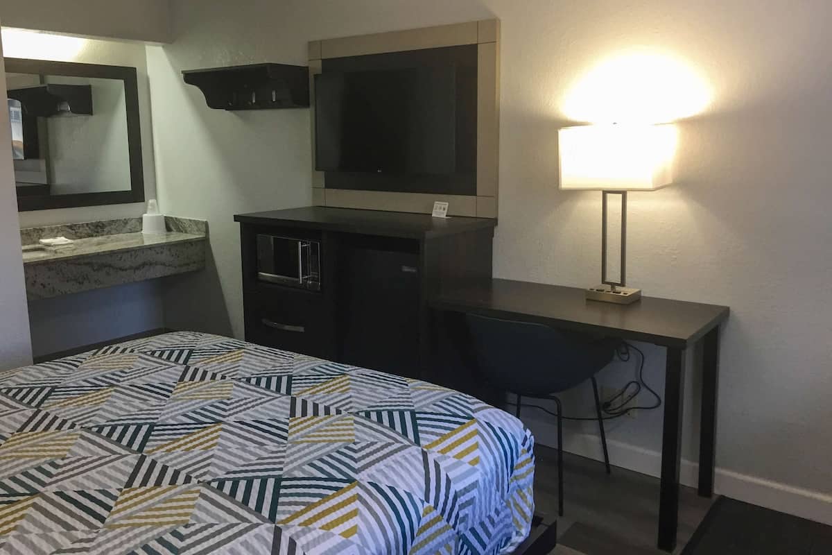 A hotel room with a bed, a desk, a chair, a mirror, and a lamp.