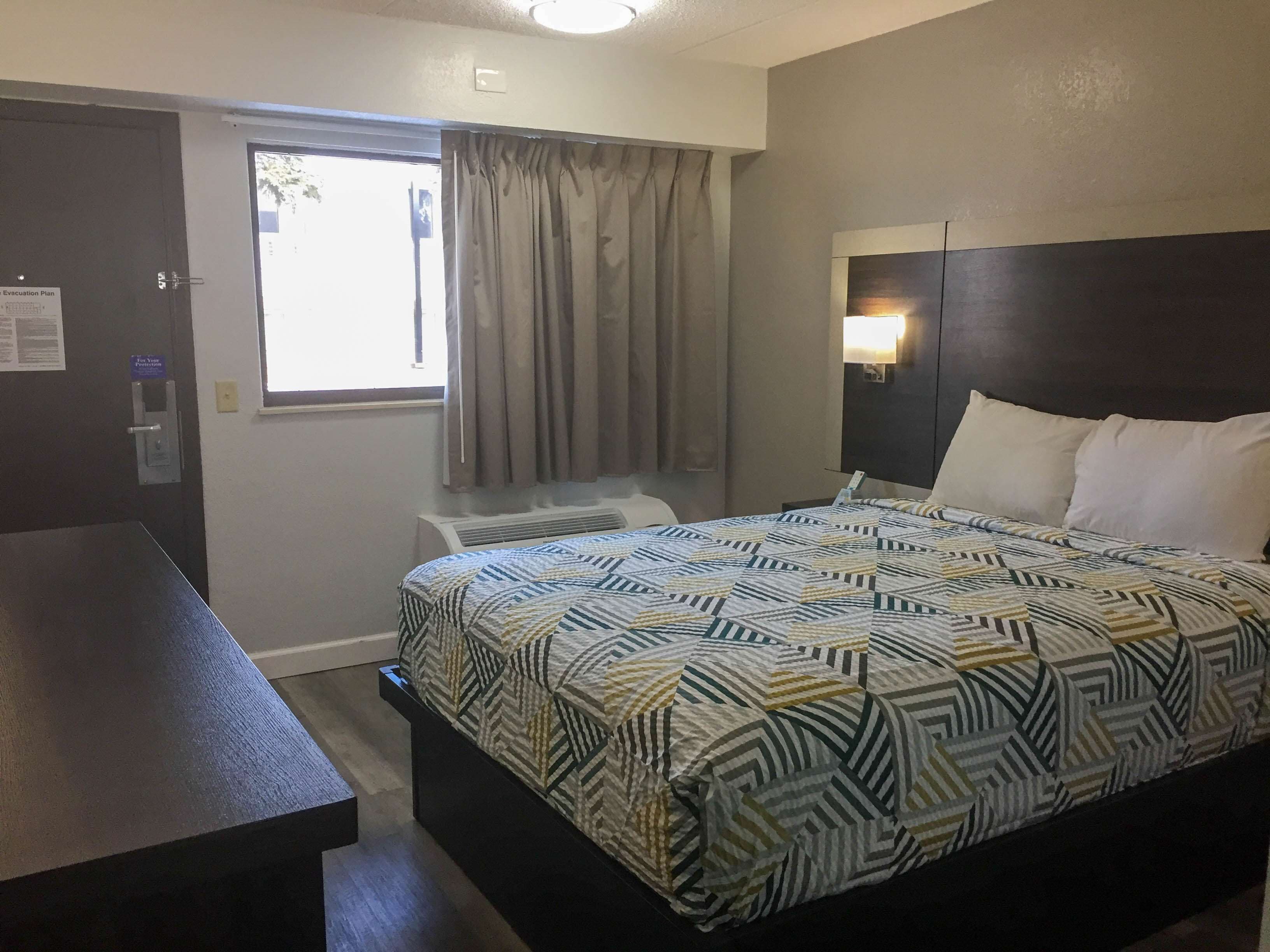 A hotel room with a bed, a desk, a window with curtains, and a wall-mounted light.