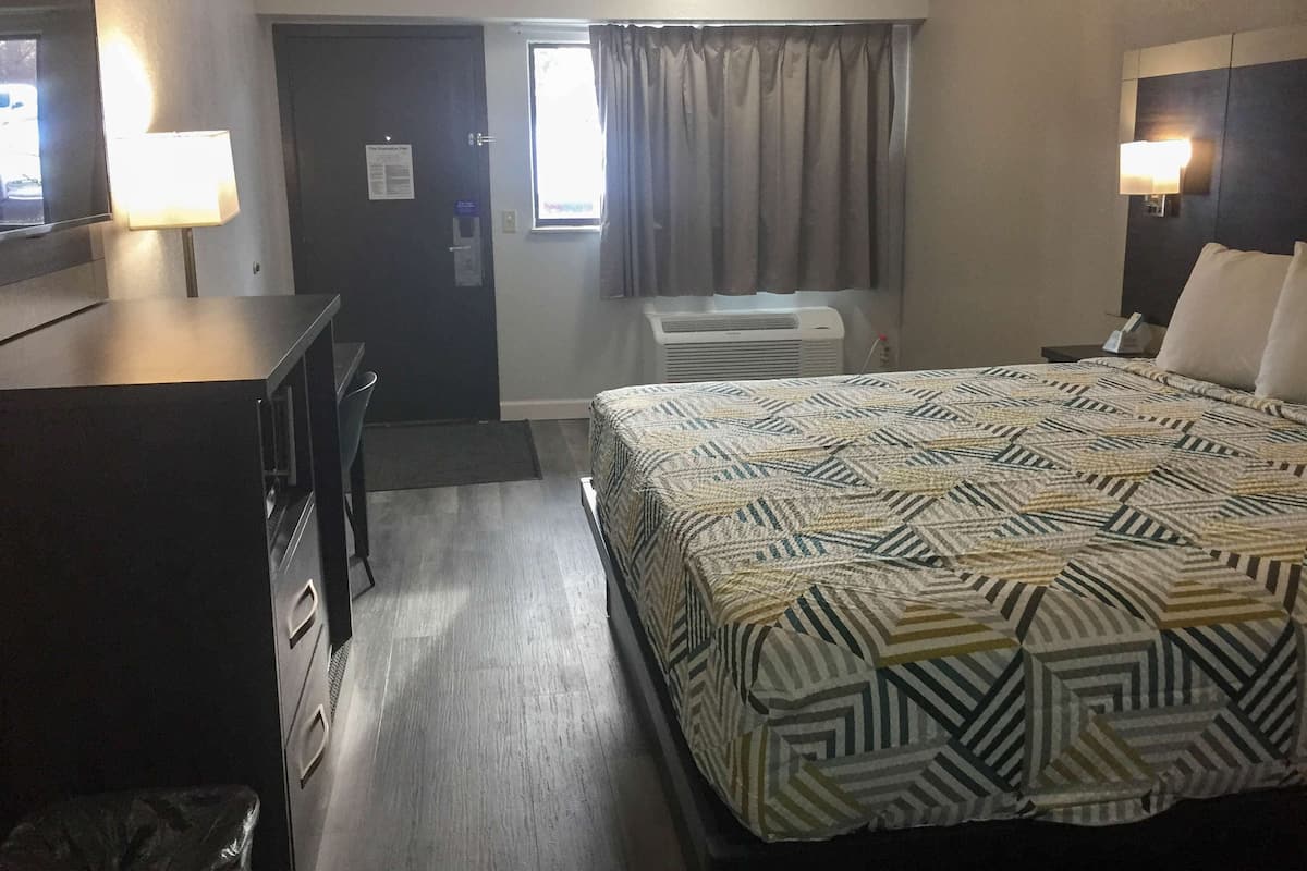 A hotel room with a bed, desk, and dresser. There is a window with curtains, an air conditioning unit, and a door with a sign.