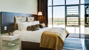 Una habitación de hotel moderna con una cama grande, una mesita de noche con lámpara, una estantería de cristal y vistas al exterior a través de amplias ventanas.
