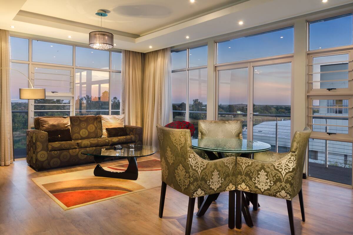 A spacious living room with a large window, a sofa, a glass coffee table, and patterned chairs.