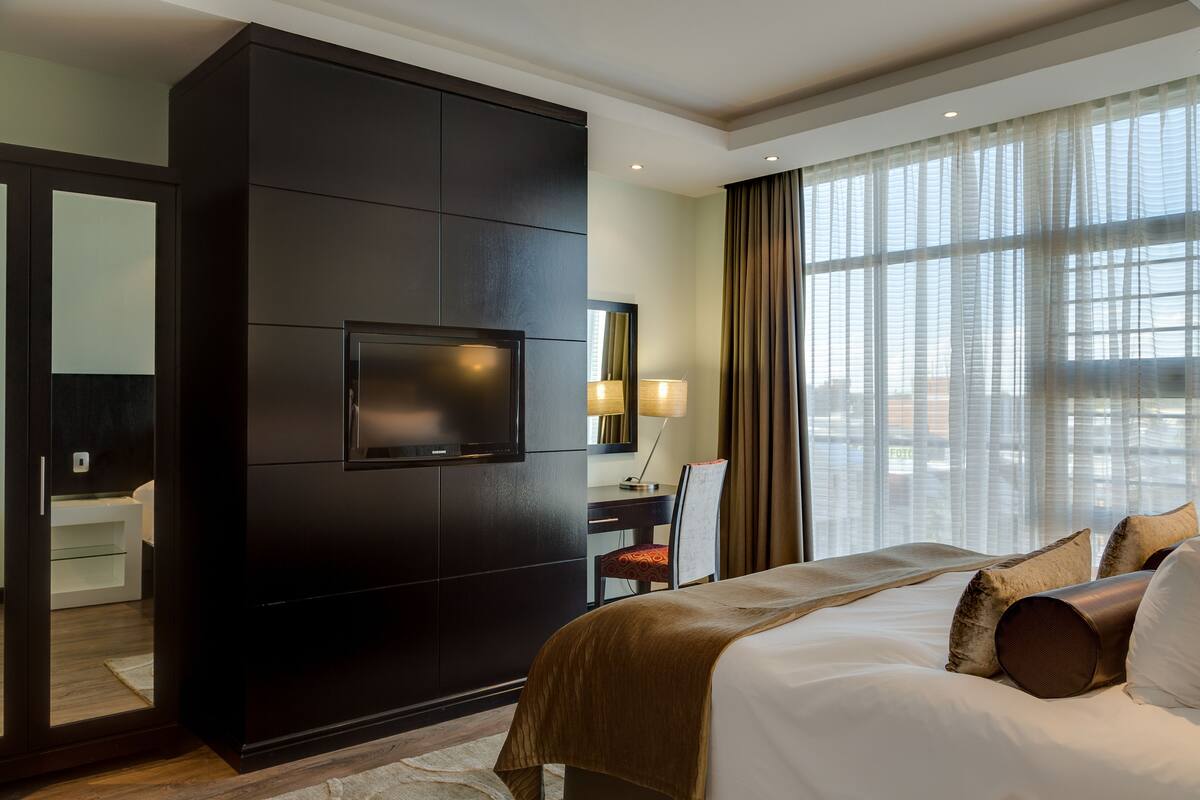 A hotel room with a large bed, a dark wooden wardrobe, a television, a desk with a chair, and a large window with curtains.