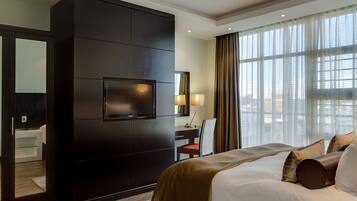 A hotel room with a large bed, a dark wooden wardrobe, a television, a desk with a chair, and a large window with curtains.