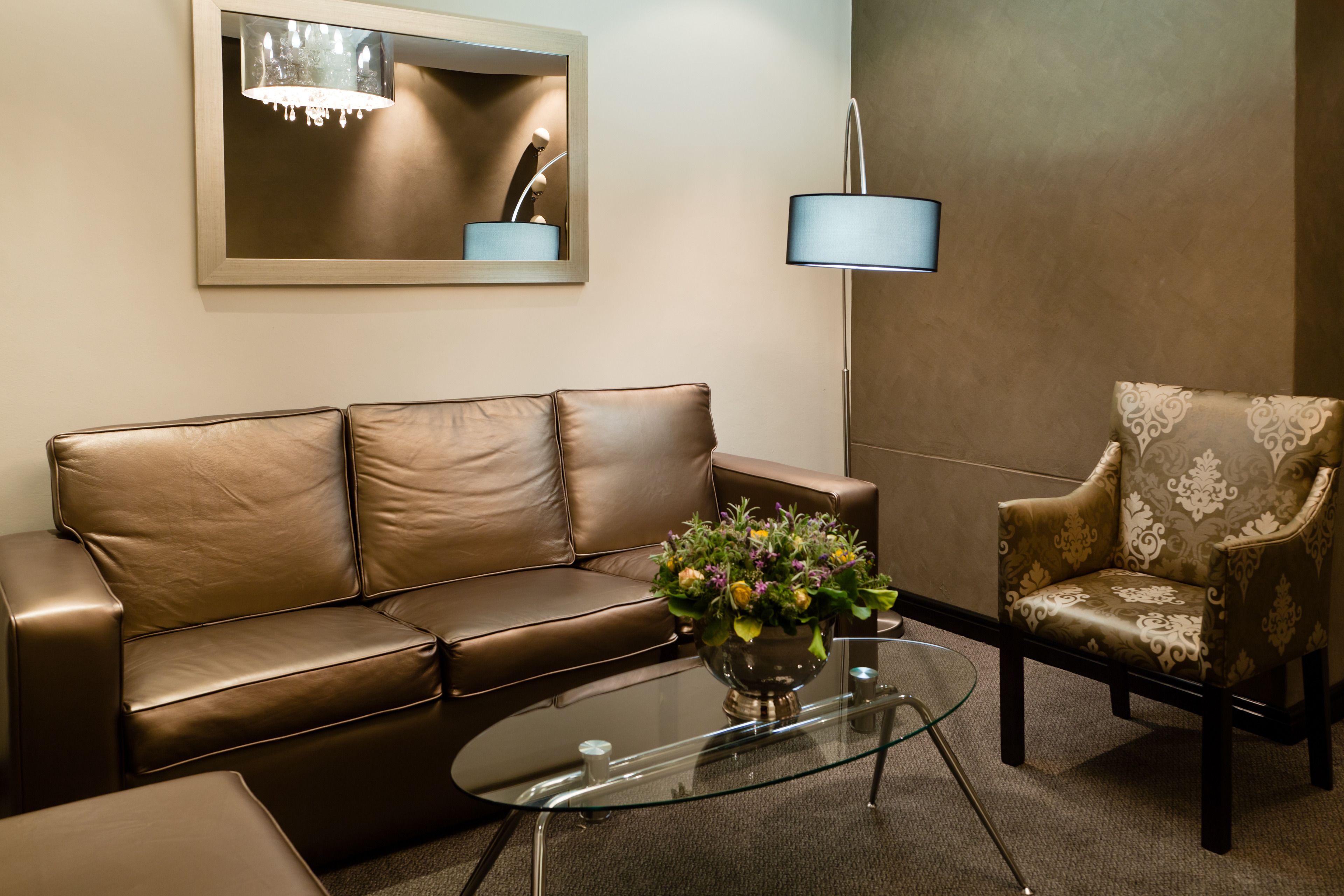 A brown leather sofa, a patterned armchair, a glass coffee table with a floral centerpiece, a floor lamp, and a mirror on the wall.