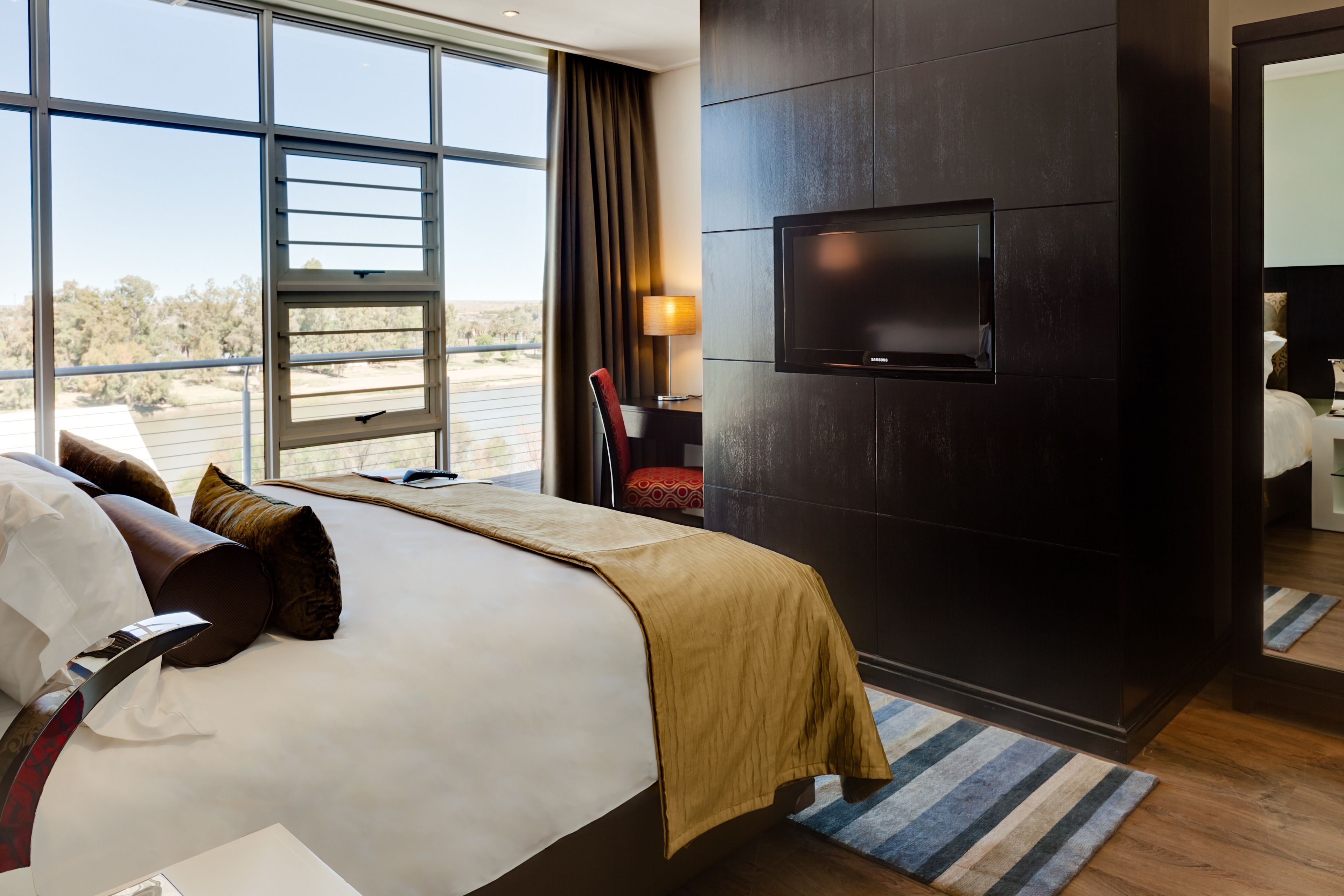 A modern hotel room with a large bed, a flat-screen TV mounted on the wall, and a view of the desert landscape through the window.