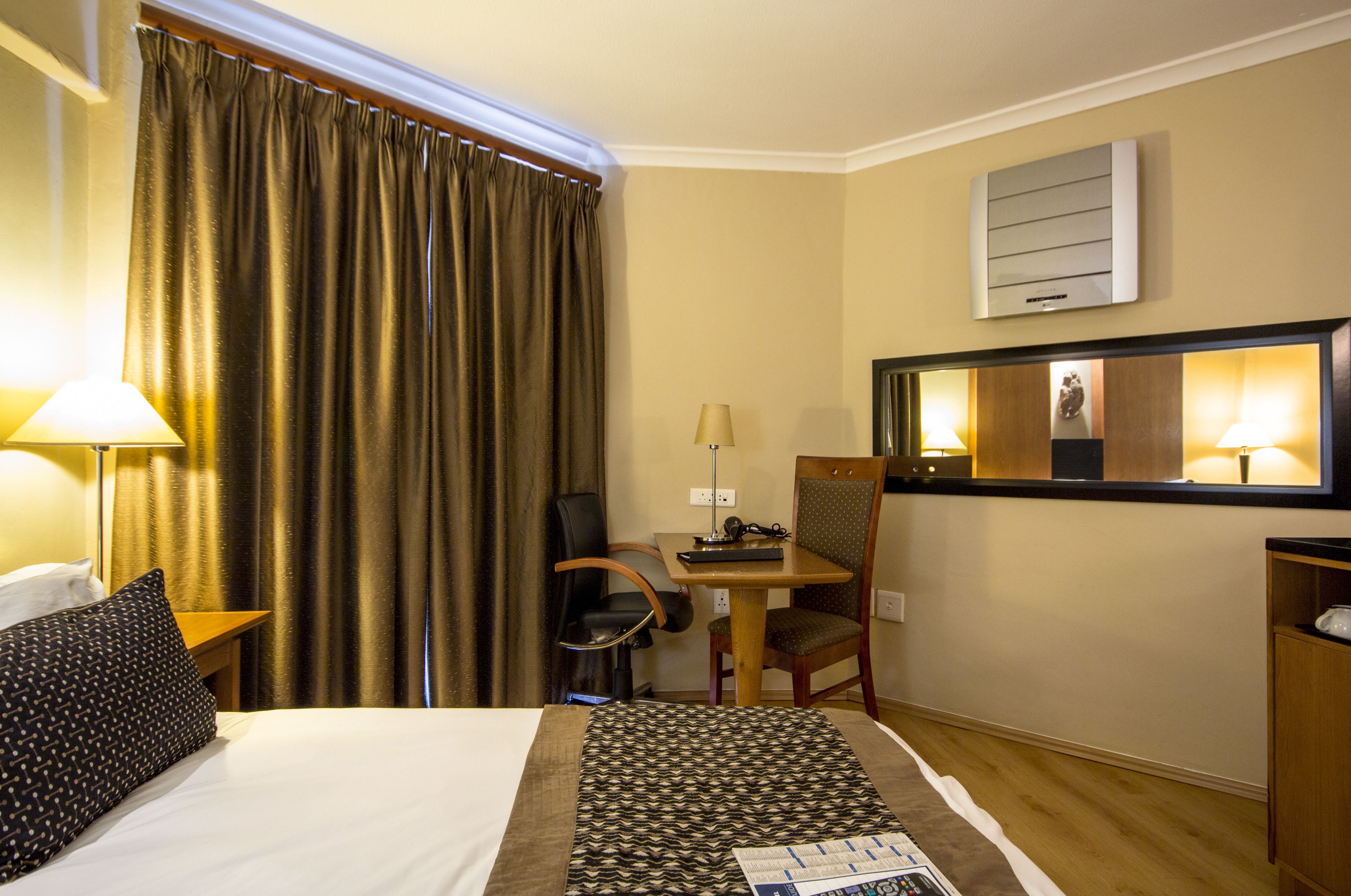 A hotel room with a bed, a desk, a chair, a lamp, a mirror, and a window with curtains.