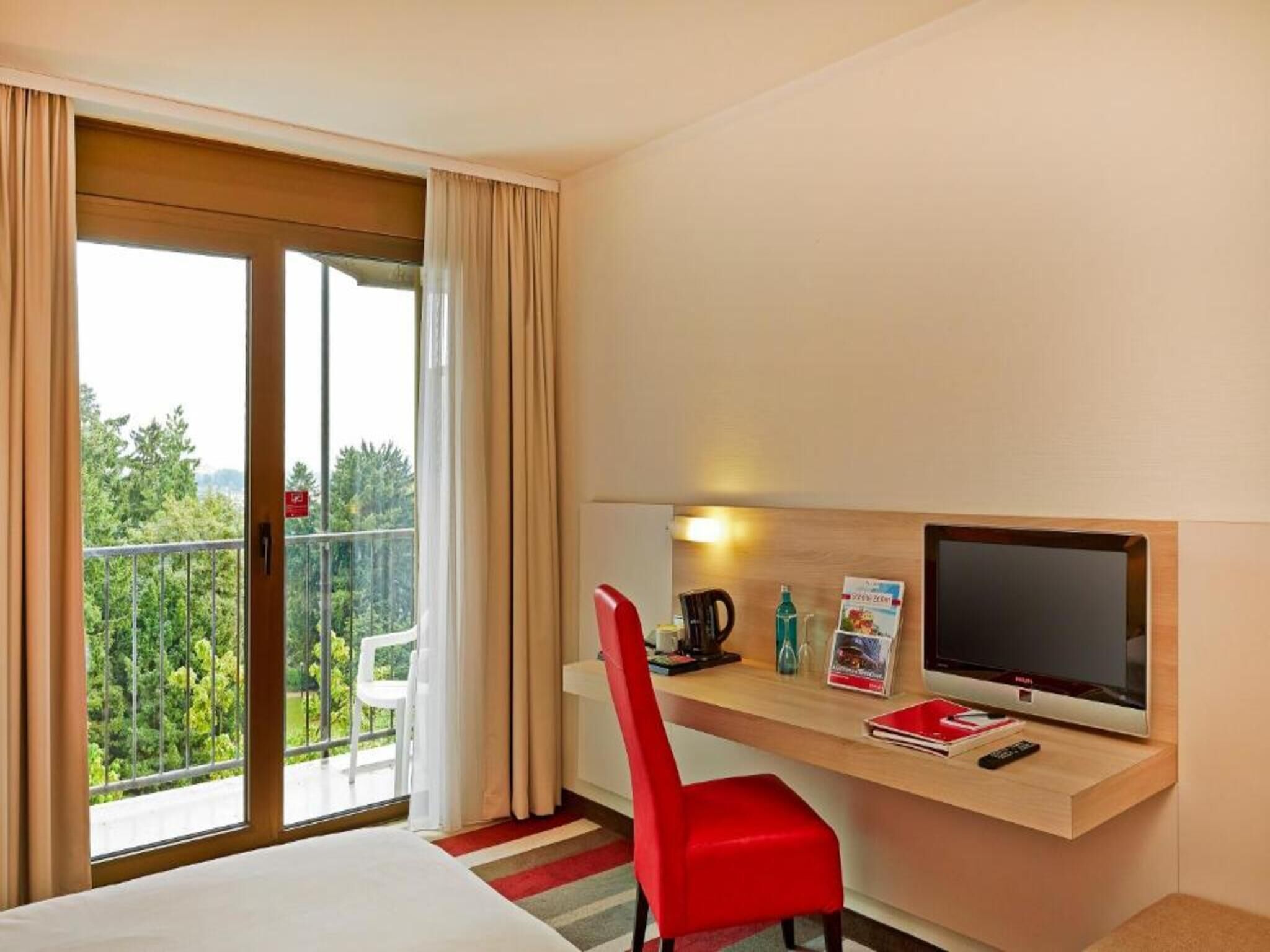 Una habitación de hotel con cama, un escritorio con televisor, una silla roja y vistas a árboles por la ventana.