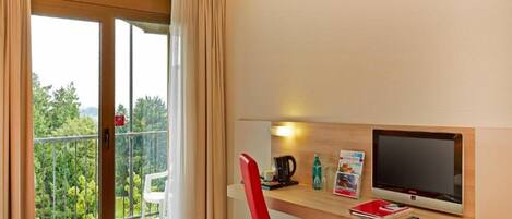 Una habitación de hotel con cama, un escritorio con televisor, una silla roja y vistas a árboles por la ventana.